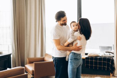 Anne bebeğini öperken, baba onu sevgiyle kucaklıyor ve karısını doğal ışıkla dolu, modern, rahat bir oturma odasında kucaklıyor, sıcak bir aile sevgisi ve birliktelik ortamı yaratıyor.