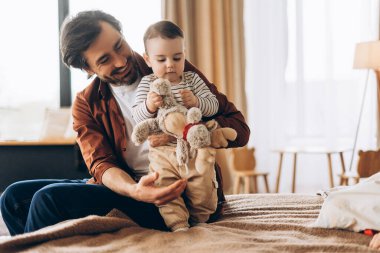 Mutlu bir baba, sıcak bir yatak odasında yatağında oturup birlikte geçirdikleri keyifli zamanların tadını çıkarırken oyuncak ayıyla oynayan oğluna sarılıp bakıyor. Aile kavramı