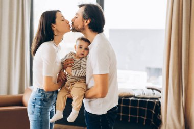 Sevecen ebeveynler sevimli bebeklerini kucaklarken şefkatli bir öpüşme paylaşıyorlar. Evlerindeki fotokopi alanlarının rahatlığında aile sevgisinin ve birlikteliğin iç açıcı sahnesini yaratıyorlar. Ebeveynlik kavramı