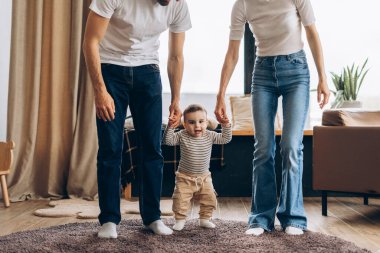 Küçük bir aile çocuklarına yürümeyi öğretiyor, ellerini tutuyor ve evdeki sıcak oturma odasında ilk adımlarını atarken neşeli anlarını paylaşıyorlar. Aile kavramı