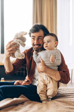 Mutlu bir baba, yatak odasındaki bir yatakta oturmuş bebeğini pelüş bir oyuncakla eğlendirirken aile bağlarını kuvvetlendiriyor. Çocukluk kavramı
