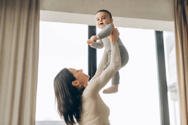 Gülümseyen anne, evdeki parlak bir pencerenin yanında neşeli bir şekilde bebeğini kaldırıyor, sevgi dolu ve neşeli bir atmosferde neşeli bir anı paylaşıyor. Annelik kavramı