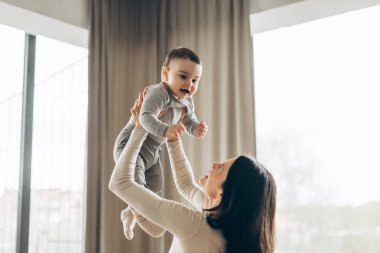 Genç anne mutlu bebeğini havaya kaldırıyor, modern oturma odalarında birlikte oynuyorlar, parlak ve havadar bir evde kaliteli aile zamanlarının tadını çıkarıyorlar. Annelik kavramı