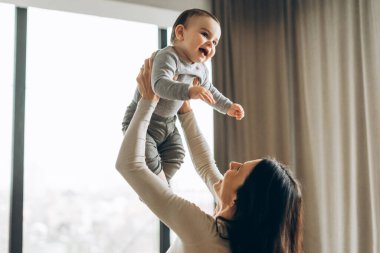 Neşeli anne, modern dairenin parlak, rahat odasında oynarken bebeğini havaya kaldırıyor. Ebeveynlik, aile, aşk ve bakım kavramı