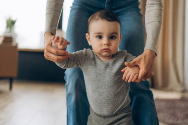 Yavrusunun sıcak evinde ilk adımlarını atmasına yardım eden genç anneyi yakından çek. Bebek kameraya bakıyor. Ebeveynlik, aile, aşk ve bakım kavramı