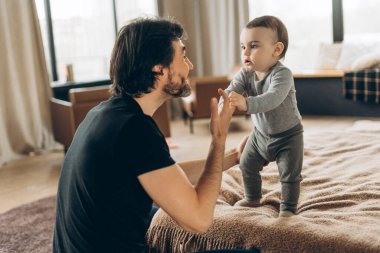 Mutlu bir babanın portresi. Evde yatak odasında ayaklarının üzerinde durmayı öğretirken minik elini nazikçe tutuyor. Ebeveynlik, aile, aşk ve bakım kavramı