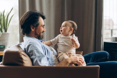 Evdeki koltukta otururken kucağında bebeğini tutan gülümseyen genç bir babanın portresi. Ebeveynlik, aile, aşk ve bakım kavramı