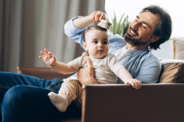 Mutlu, genç bir baba, evde yan yana otururken oğlunun saçlarını nazikçe tarıyor. Ebeveynlik, aile, aşk ve bakım kavramı