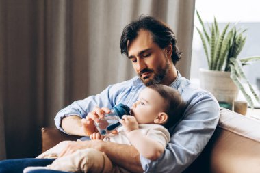 Çocuk şişeden su içerken küçük oğlunu kucağında tutan mutlu genç babanın portresi. Ebeveynlik, aile, aşk ve bakım kavramı