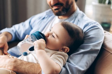 Gülümseyen neşeli genç babaya yakın çekim, evdeki su yedirme sırasında bebeğine destek oluyor. Ebeveynlik, aile, aşk ve bakım kavramı