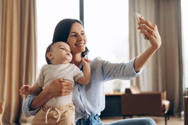 Küçük oğlunu kucağında tutan gülümseyen bir annenin portresi ve akıllı telefon kullanarak evde selfie çekerken. Ebeveynlik, aile, aşk ve bakım kavramı