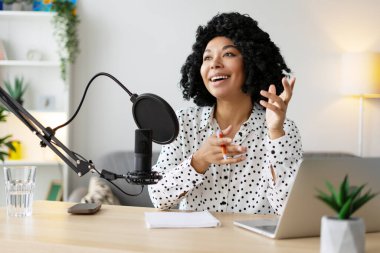 Neşeli kadın podcaster evde ses kaydı yaparken mikrofonla meşgul oluyor, modern, rahat bir stüdyo ortamında içerik yaratıyor.