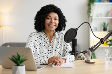 Mutlu kadın podcaster mikrofona konuşuyor, masasında otururken yaratıcı aletlerle çevrili bir ses yayını kaydediyor.