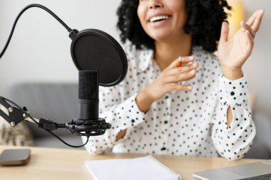 Neşeli kadın podcaster ses içeriğini kaydediyor, bir mikrofona konuşuyor ve stüdyosunda otururken el kol hareketi yapıyor.