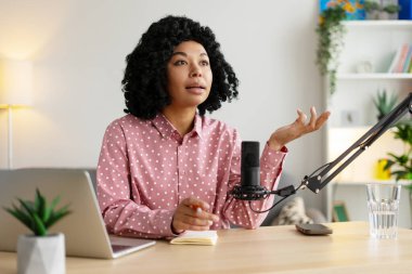 Ev stüdyosundan podcast yapan genç bir kadın mikrofona konuşuyor ve yukarı bakıyor, not defterine notlar alıyor.