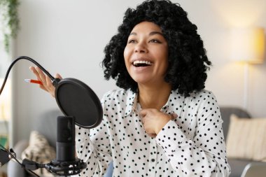 Neşeli kadın podcaster yeni bir bölüm kaydederken gülüyor, ses ekipmanlarıyla dolu rahat stüdyosundaki yaratıcı sürecin tadını çıkarıyor.