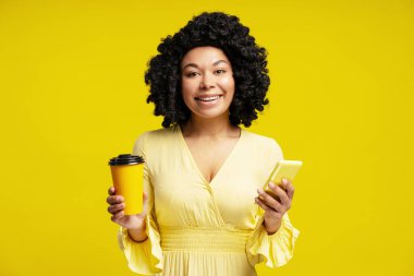 Kahveden zevk alan ve akıllı telefonunu kullanan mutlu bir kadın, parlak sarı bir arka planda izole edilmiş.