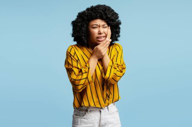 Kıvırcık saçlı, sarı çizgili tişörtlü, mavi arka planda diş ağrısı çeken genç bir kadının stüdyo fotoğrafı.