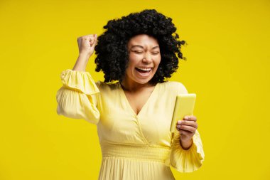 Aşırı neşeli Afro-Amerikalı kadın akıllı telefonuna bakarken elini kaldırıyor, online zaferi kutluyor ya da sarı arka planda iyi haberler var.