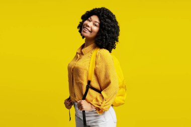 Sarı gömlek ve sırt çantası giyen, sarı arka planda poz veren neşeli genç bir öğrencinin portresi.