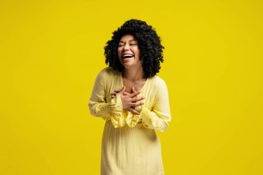 Canlı sarı bir arka planda, elleri göğsünde gülen neşeli genç bir kadının stüdyo portresi.
