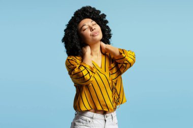Stüdyo portresi kıvırcık saçlı, sarı çizgili bluz ve beyaz kot giyen, zevk veren ve rahatlatan, açık mavi arka planda boynuna dokunan genç bir kadın.