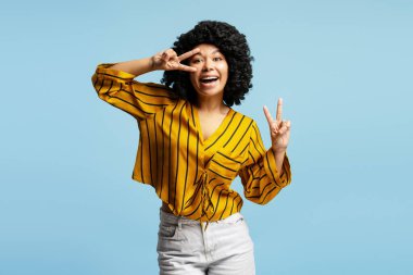 Mavi stüdyo arka planında izole edilmiş, iki eliyle barış işareti yapan ve kameraya gülümseyen neşeli genç bir kadının portresi.