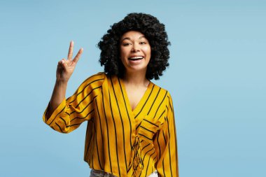 Neşeli genç bir kadın, gülümsüyor ve parmaklarıyla bir barış işareti yapıyor. Parlak mavi bir arka plan, neşe ve pozitiflik saçıyor.