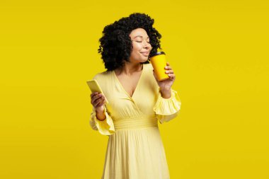 Stüdyo portresi, elinde akıllı bir telefon tutan ve sarı arka planda kapalı gözleri olan bir fincan kahveyi koklayan genç bir kadın.