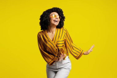 Kıvırcık saçlı, sarı gözlüklü ve sarı arka planda çizgili gömlekli neşeli genç bir kadının stüdyo portresi.