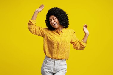 Kaygısız genç kadın dans ediyor ve kapalı gözlerle şarkı söylüyor, müzikten hoşlanıyor ve canlı sarı bir arka planda olumlu duygular ifade ediyor.