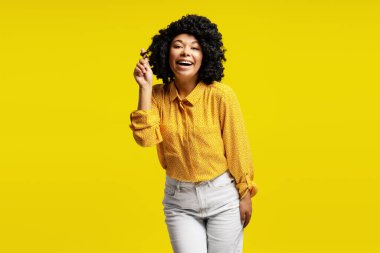 Kıvırcık saçlı neşeli kadın saçlarıyla oynuyor ve canlı sarı bir arka planda gülümsüyor.