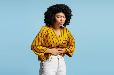 Stüdyo çekiminde kıvırcık saçlı, çizgili bluzlu, güçlü bir karın ağrısı çeken, mavi arka planda izole edilmiş genç bir kadın görülüyor.