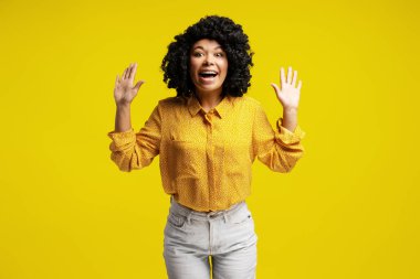 Heyecanlı, kıvırcık saçlı ve sarı gömlekli genç bir kadın heyecanlı sarı arka planda el kaldırıyor, neşe ve coşkuyu ifade ediyor.