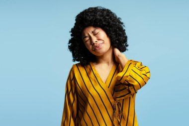 Boynu ağrıyan genç bir kadının stüdyo portresi mavi arka planda acı veren bir ifadeyle boynuna dokunuyor.