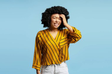 Alnına dokunan kıvırcık saçlı ve sarı çizgili tişörtlü genç bir kadının stüdyo portresi. Mavi arka planda izole edilmiş şiddetli bir baş ağrısından muzdarip.