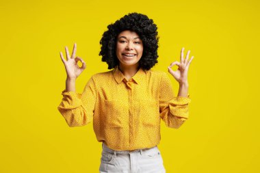 Gülümseyen kadın her iki eliyle iyi bir jest yaparken canlı sarı bir arka plana karşı duruyor, pozitiflik ve güven yayıyor.