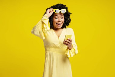 Heyecanlı genç kadın sarı güneş gözlüğünü kaldırıyor ve sarı arka planda akıllı telefonundan iyi haberler okuyor.