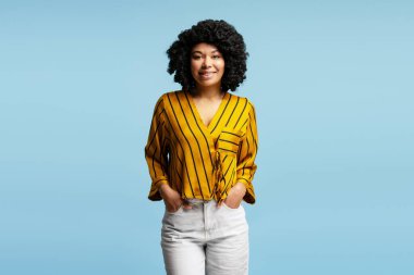 Sıradan kıyafetler giyen, elleri cebinde, mavi arka planda gülümseyen genç bir kadının stüdyo portresi.