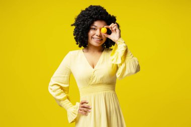 Sarı elbiseli neşeli genç bir kadının atölye çekimi. Gözünün üstüne küçük bir top tutuyordu. Parlak sarı bir zemine karşı neşeli bir enerji saçıyordu.