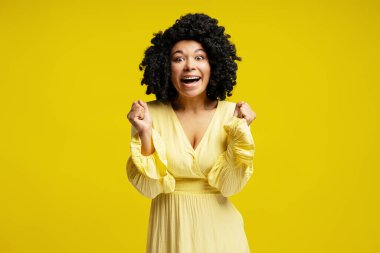 Kıvırcık saçlı, sarı elbiseli, heyecanlı genç bir kadın. Başarımızı canlı sarı bir arka planda yumruklarımızla kutluyoruz.