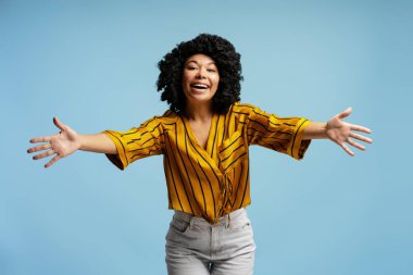 Heyecanlı kıvırcık saçlı, sarı çizgili tişörtlü, kollarını açıp kucaklaşan açık mavi arka planda sevgi ve destek sunan genç bir kadın.