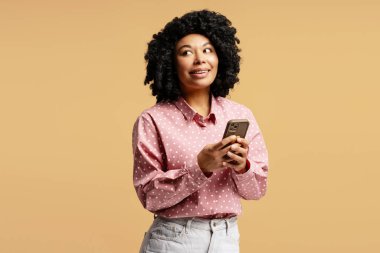 Kıvırcık saçlı, pembe tişörtlü, elinde akıllı telefon olan ve bej arka planda gülümseyen genç bir kadın.