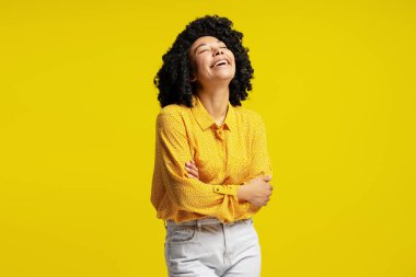 Kollarını kavuşturmuş neşeli genç bir kadının parlak sarı bir arka plana gülüşü neşe ve mutluluğu ifade eden stüdyo portresi.