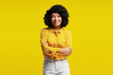 Sarı gömlekli, beyaz pantolonlu, sarı arka planda poz veren çapraz kollu neşeli genç bir kadının stüdyo portresi.