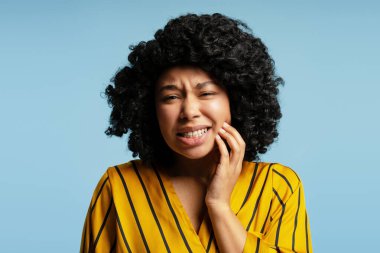 Kıvırcık saçlı, sarı ve siyah çizgili gömlekli mavi arka planda diş ağrısı olan genç bir kadının stüdyo portresi.