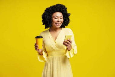 Cep telefonu kullanan ve kahve fincanını sarı arka planda tutan gülümseyen kadının stüdyo portresi.