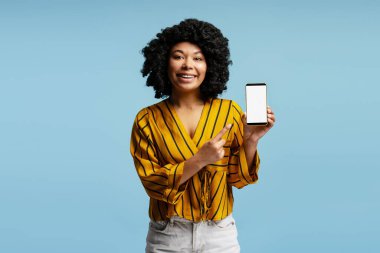 Beyaz ekranlı akıllı telefonu gösteren heyecanlı genç bir kadın, cep telefonu ekranını işaret ediyor, başvuru, web sitesi veya reklam öneriyor, mavi stüdyo arka planında poz veriyor.