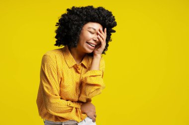Kıvırcık saçlı, neşeli genç bir kadının stüdyo portresi. Gülümseyen ve elini parlak sarı bir arka planda tutarak yüzünü kapatan bir kadın.