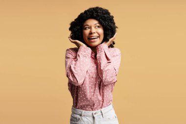 Elleriyle yanaklarını tutarak bej bir arka plana bakan kıvırcık saçlı, şaşırmış genç bir kadının stüdyo portresi.